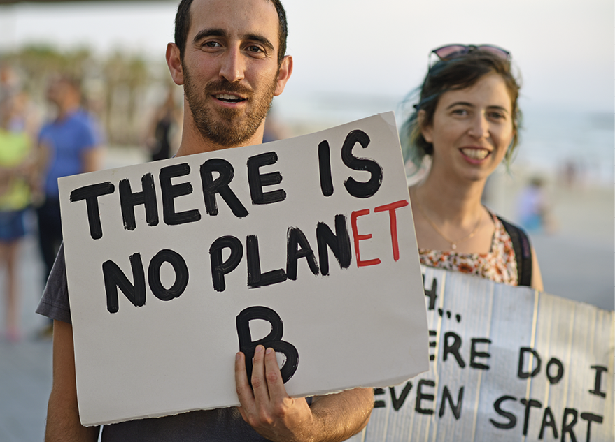 Fotografia. À esquerda, um homem visto da cintura para cima, de cabelos e barba rala em preto, com camiseta cinza e segurando na mão direita, um cartaz branco com texto preto: There is no planet B. Mais atrás, uma mulher, de cabelos escuros, com par de óculos de sol sobre a cabeça, blusa clara, mochila preta sobre as cotas. Ela olha para frente sorrindo e segurando um cartaz nas mãos, com texto ilegível.