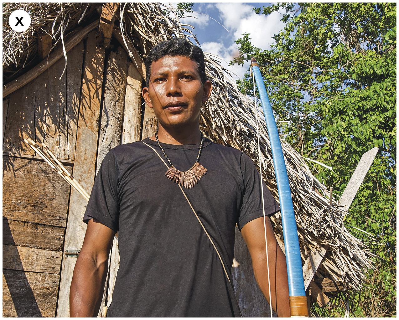 Fotografia. Número dez. Um homem indígena visto de baixo para cima da cintura até a cabeça. Ele tem cabelos pretos curtos e ondulados. Está usando camiseta preta e colar marrom, olhando para a frente e sorrindo suavemente. Ele segura um arco azul na mão esquerda e nas costas carrega algumas flechas. Ao fundo, vista parcial de casa de madeira com telhado de palha. À direita, árvore de folhas verdes. No alto, céu em azul-claro e nuvens em branco.