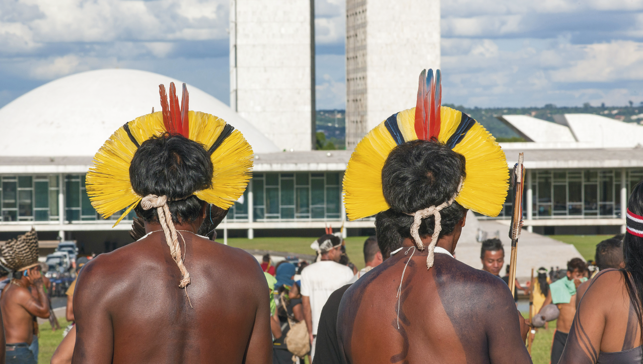 Fotografia. À frente, dois homens indígenas vistos de costas, da cintura para cima. Eles têm cabelos pretos, com cocar sobre a cabeça em amarelo, partes em preto e partes em vermelho. Ao fundo, outras pessoas vistas parcialmente. Mais ao fundo, prédio do Congresso Nacional do Brasil em Brasília, um edifício composto por duas torres gêmeas, uma delas mais alta que a outra, interligadas por uma estrutura em forma de arco com aparência esculpida, com curvas suaves e elegantes. As duas torres, também feitas de concreto, têm uma aparência sólida e retangular. No alto, céu em azul-claro com nuvens brancas.