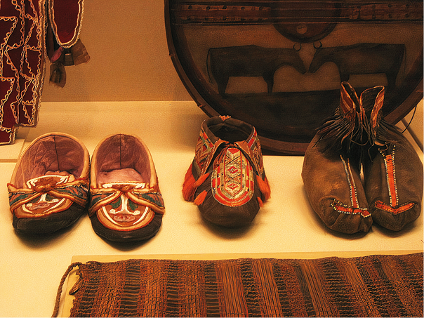 Fotografia. Sapatos feitos com tecido escuro com algumas partes coloridas. Um dos sapatos é mais baixo. O sapato do meio tem um cano intermediário e o sapato da direita tem o cano alto com franjas nas laterais.