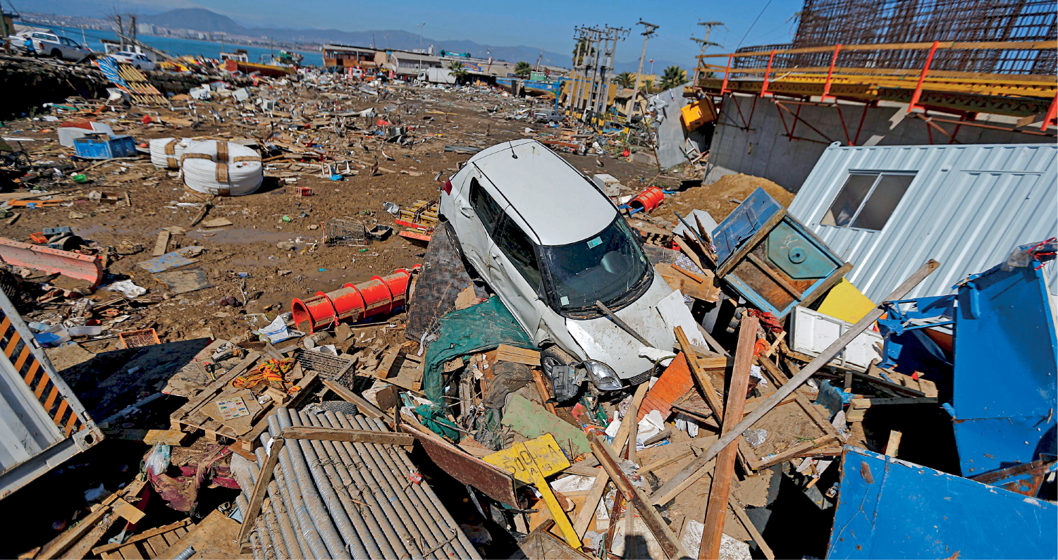 Fotografia. Destroços espalhados por uma região litorânea. À frente, grande acumulado de pedaços de madeira e outros objetos. Entre eles um carro amassado.