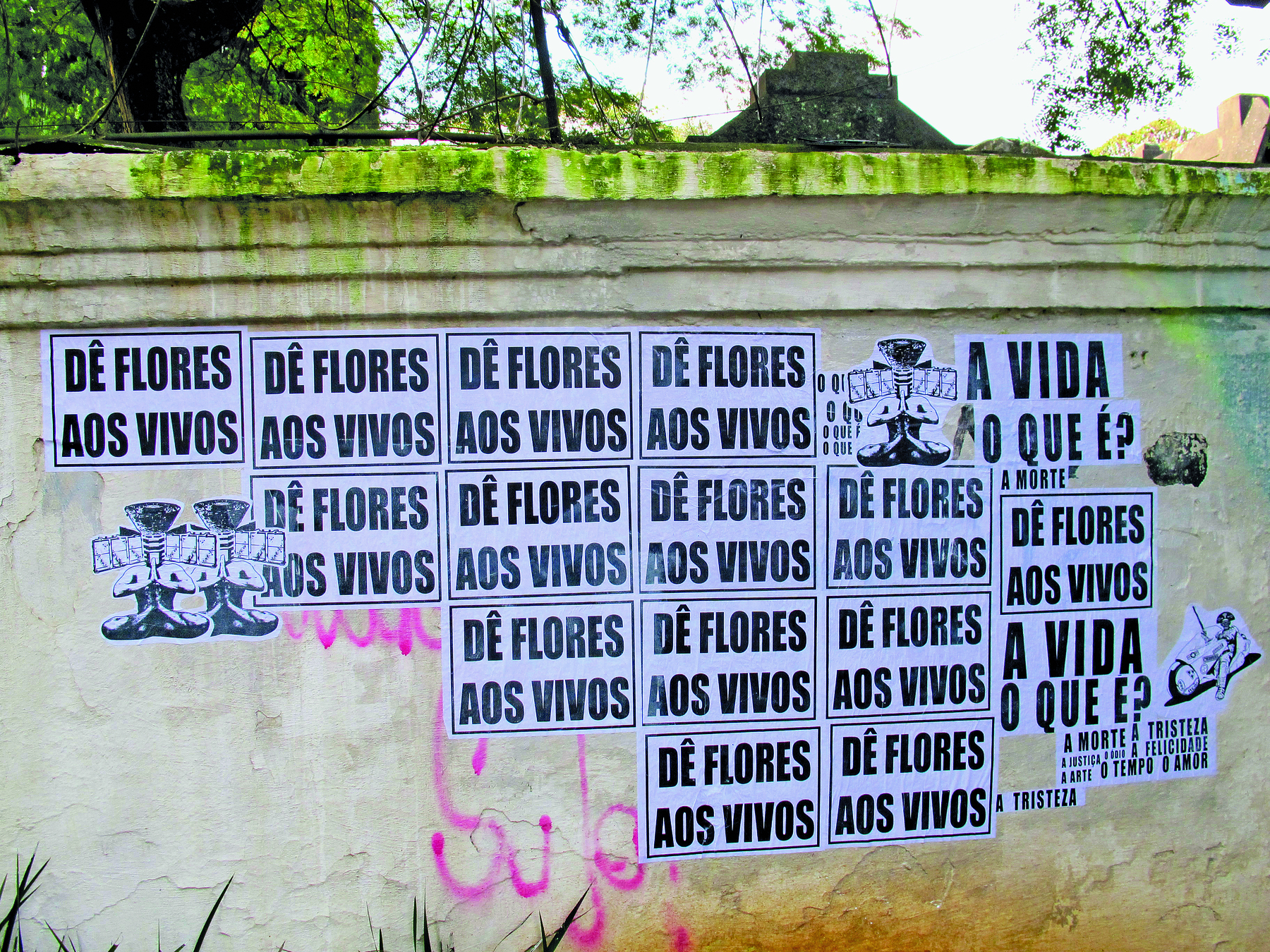 Fotografia. Muro com manchas de musgo e outras manchas. Sobre ele, diversos papéis colados. Na maioria deles está a frase: DÊ FLORES AOS VIVOS. Alguns dos papéis tem a seguinte pergunta: A VIDA O QUE É?