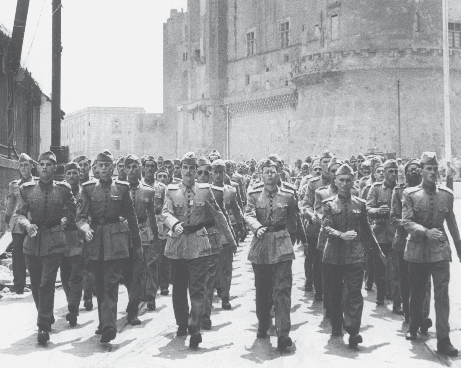 Estilo padrão;Fotografia em preto e branco. Soldados em filas marchando. Usam uniforme e quepe. Ao fundo, grandes construções.