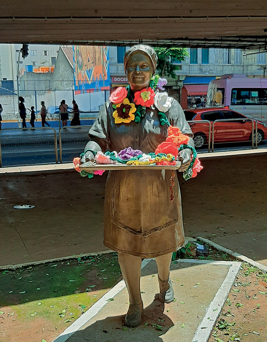 Fotografia. Estátua de uma mulher de touca, usando um vestido de uniforme. Ela carrega à frente do corpo uma bandeja. Flores de crochê estão enfeitando seu pescoço, braços e estão sobre a bandeja.