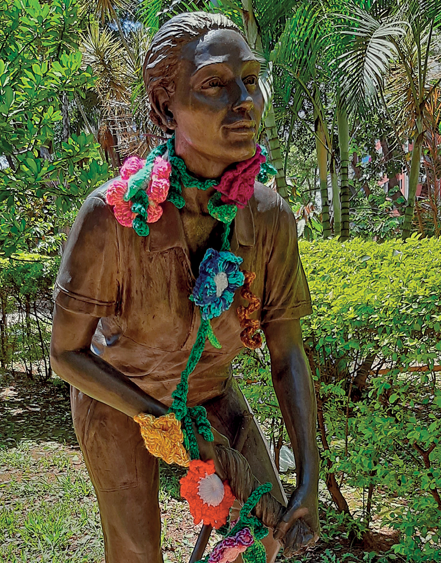 Fotografia. Estátua de um homem de uniforme, como o corpo inclinado para frente torcendo um pano com as duas mãos. Ele tem flores de crochê enfeitando seu pescoço e braço.