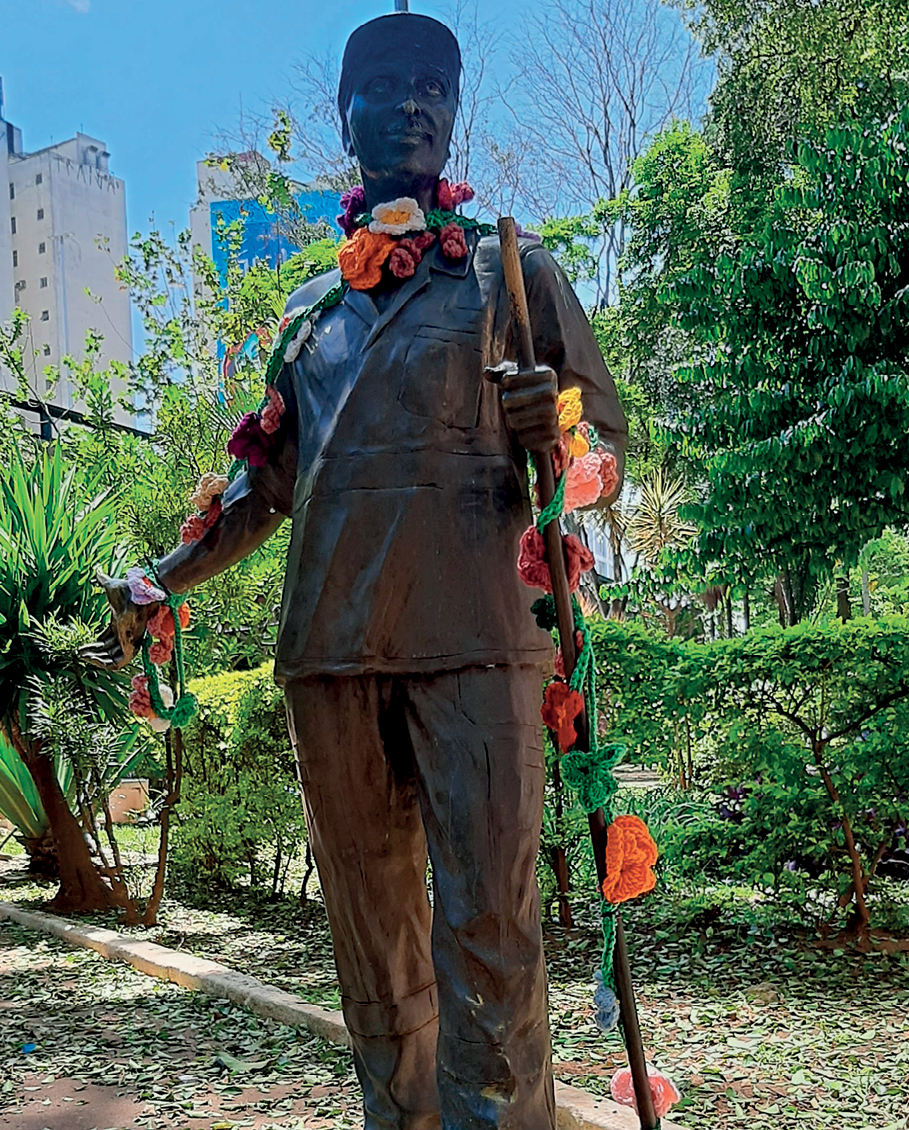 Fotografia. Estátua de um homem de boné e uniforme. Uma das mãos está aberta para o lado e na outra ele segura um cabo. Tem flores de crochê enfeitando seus braços, pescoço e o cabo.