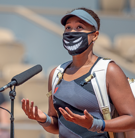 Fotografia. Naomi Osaka, atleta de cabelo curto. Usa uma viseira e máscara de proteção facial. Tem uma mochila nas costas. À frente dela um microfone.