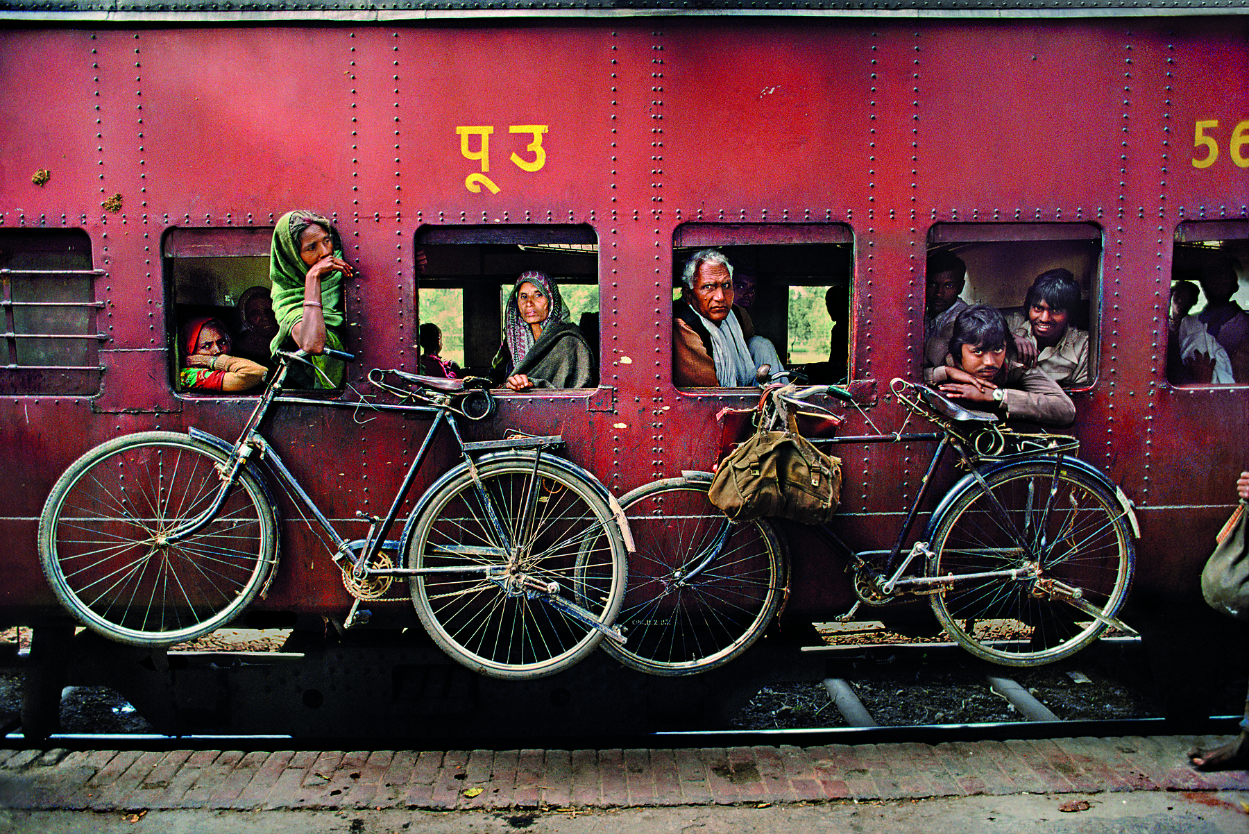 Fotografia. Pessoas dentro de um trem vermelho com janelas abertas. Do lado de fora, duas bicicletas estão penduradas. Algumas pessoas se projetam levemente para fora das janelas.