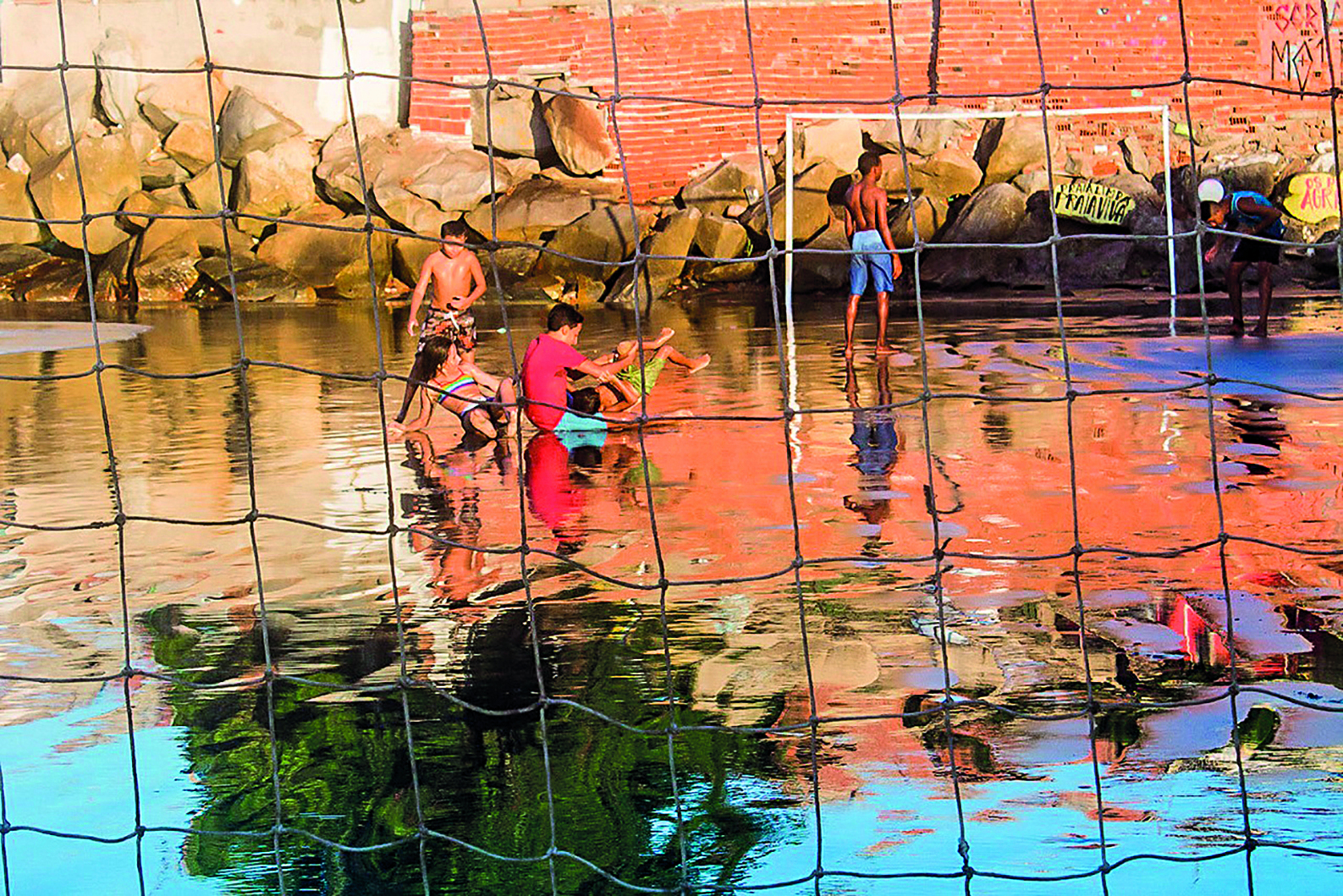 Fotografia. Crianças brincando em uma rua alagada. Na água, é possível observar o reflexo de uma árvore grande. Ao fundo, encostadas em um muro de tijolos, muitas pedras grandes. Toda a paisagem está encoberta por uma rede quadriculada.