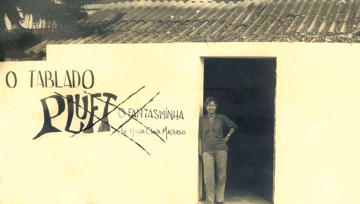 Fotografia em preto e branco. Maria Clara Machado parada na porta de uma construção térrea e telhado triangular. No muro claro, as informações:  O TABLADO PLUFT, O FANTASMINHA, de Maria Clara Machado.