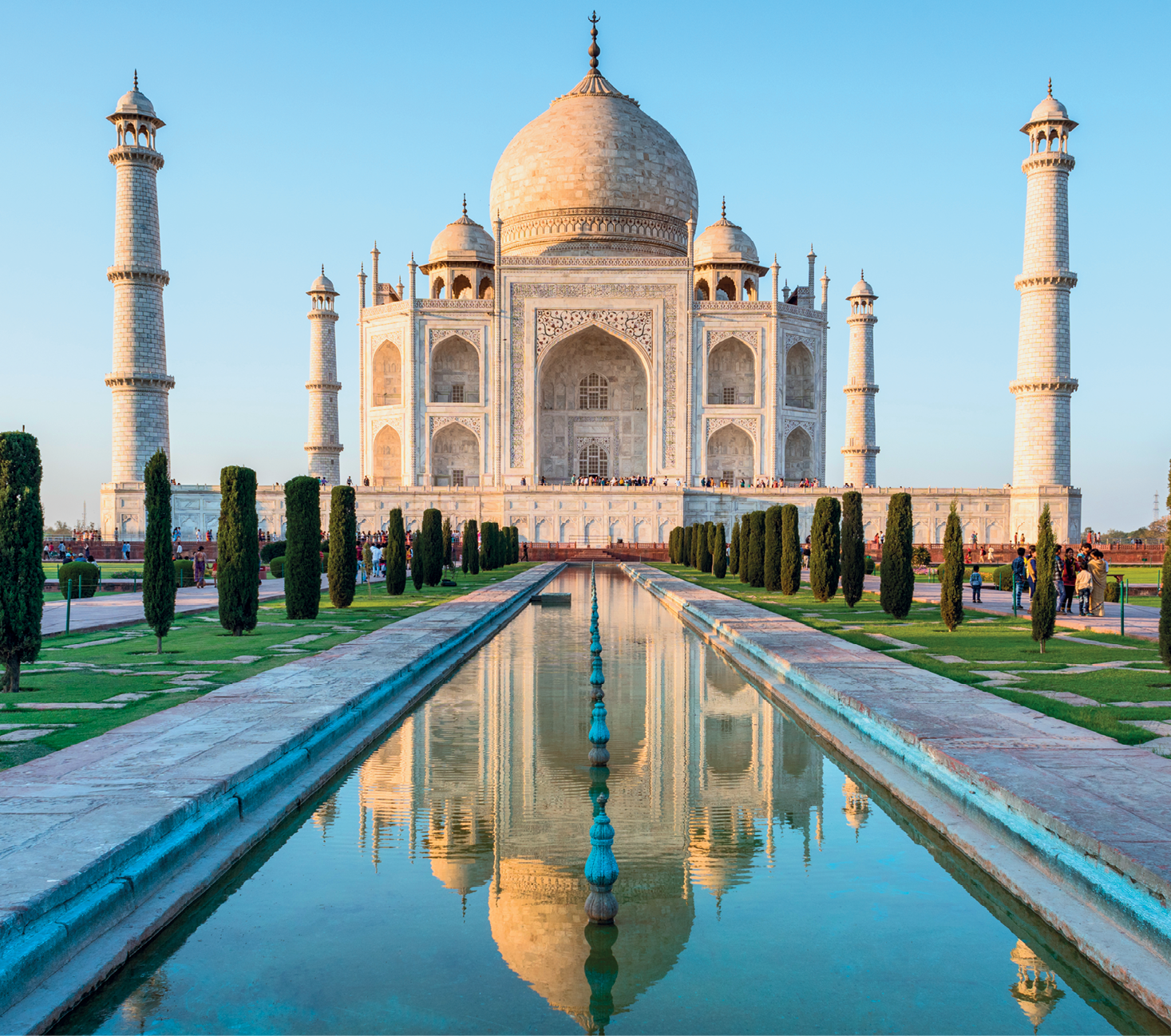 Fotografia. Taj Mahal, grande palácio de estrutura clara, com três cúpulas no topo. A central é a maior. As janelas são grandes e, ao redor delas, há aberturas gigantes na estrutura, em formato de arco. O palácio é cercado por uma muralha. Nas quatro pontas dela, torres compridas. À frente, um espelho d’água com árvores ao redor. Perto das árvores, estão dispostas algumas pessoas. O tamanho delas, pequeno, destoa do tamanho da estrutura do monumento. O céu, ao fundo, está azul.
