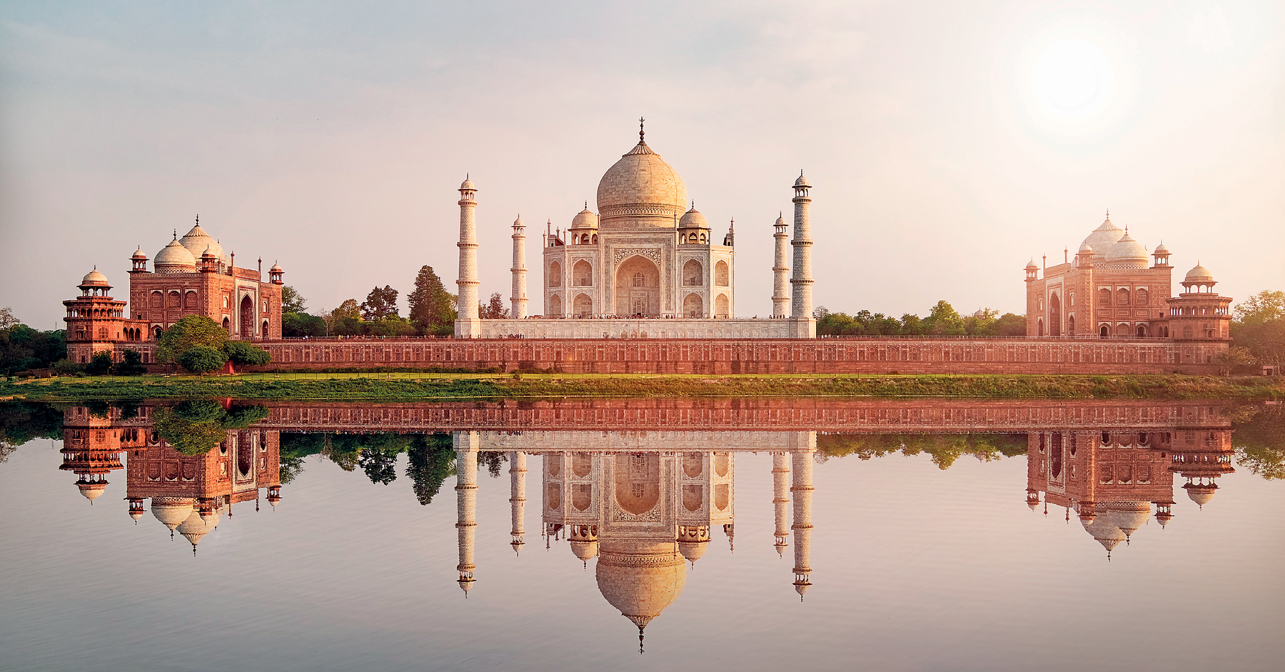 Fotografia. Taj Mahal cercado por um longo muro ornamentado. Em cada extremidade do muro, um palácio menor que o principal, com várias janelas e cúpulas arredondadas no topo. Todo o conjunto está refletido nas águas de um rio, à frente. O céu, ao fundo, apresenta tons de azul, lilás e cinza, bem claros.
