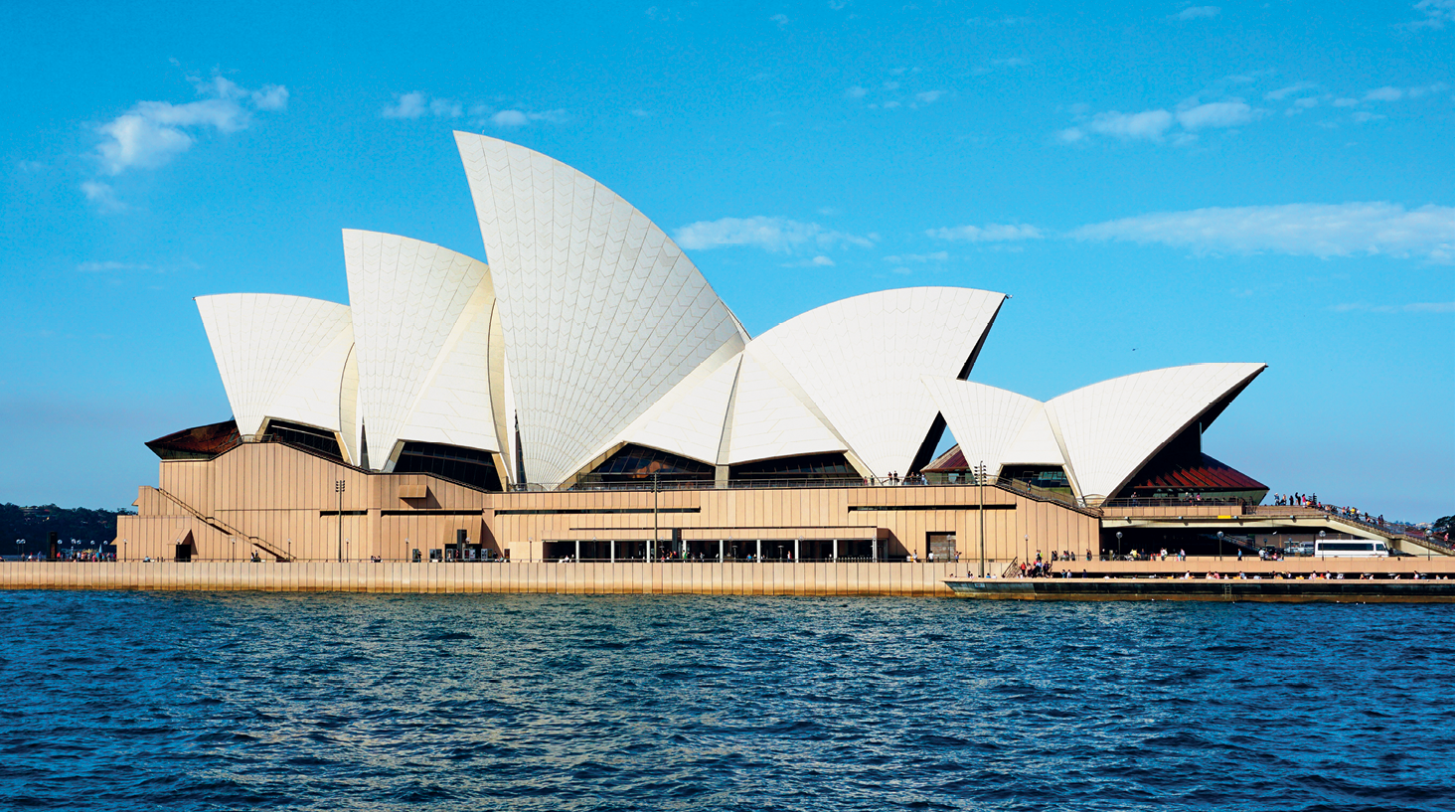 Fotografia. Casa da Ópera de Sydney, grande construção retangular à beira-mar. O telhado é branco e tem o formato de grandes conchas sobrepostas. O céu, ao fundo, está azul.