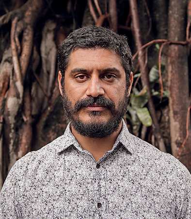 Fotografia. Criolo, homem de cabelo preto e curto. Tem barba e bigode escuros. Os olhos são pequenos e o nariz é largo. Os lábios são grandes.