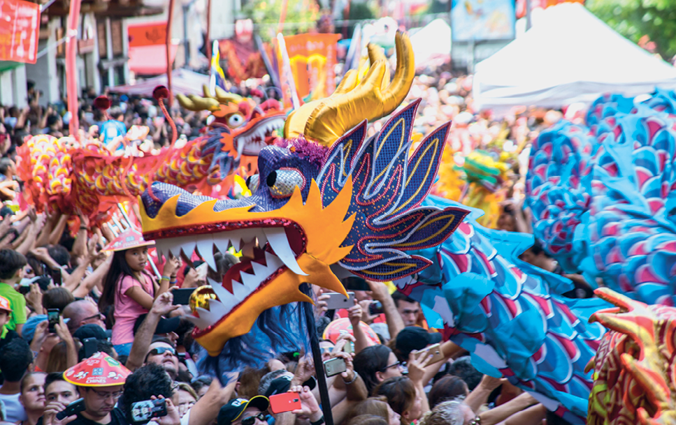 Fotografia. População aglomerada. Passando entre as pessoas, dois grandes bonecos de dragão: um azul com chifres dourados e o outro vermelho.