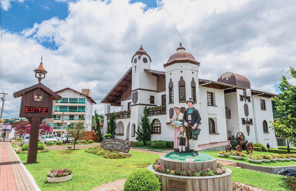 Fotografia. À frente jardim com flores e arbustos. No centro, estátua de uma família. Ao fundo, construção branca de dois andares, com muitas janelas marrom e telhado triangular, também marrom. À esquerda, um letreiro digital em uma estrutura de madeira no formato de uma casinha.