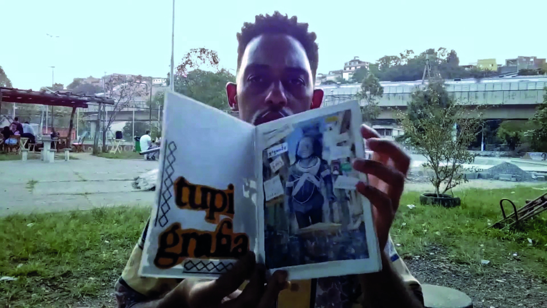 Cena de vídeo. Roger na praça com um fanzine aberto. À esquerda, um título em recortes de letras grandes:  TUPI GRAFIA. À direita, fotomontagem.