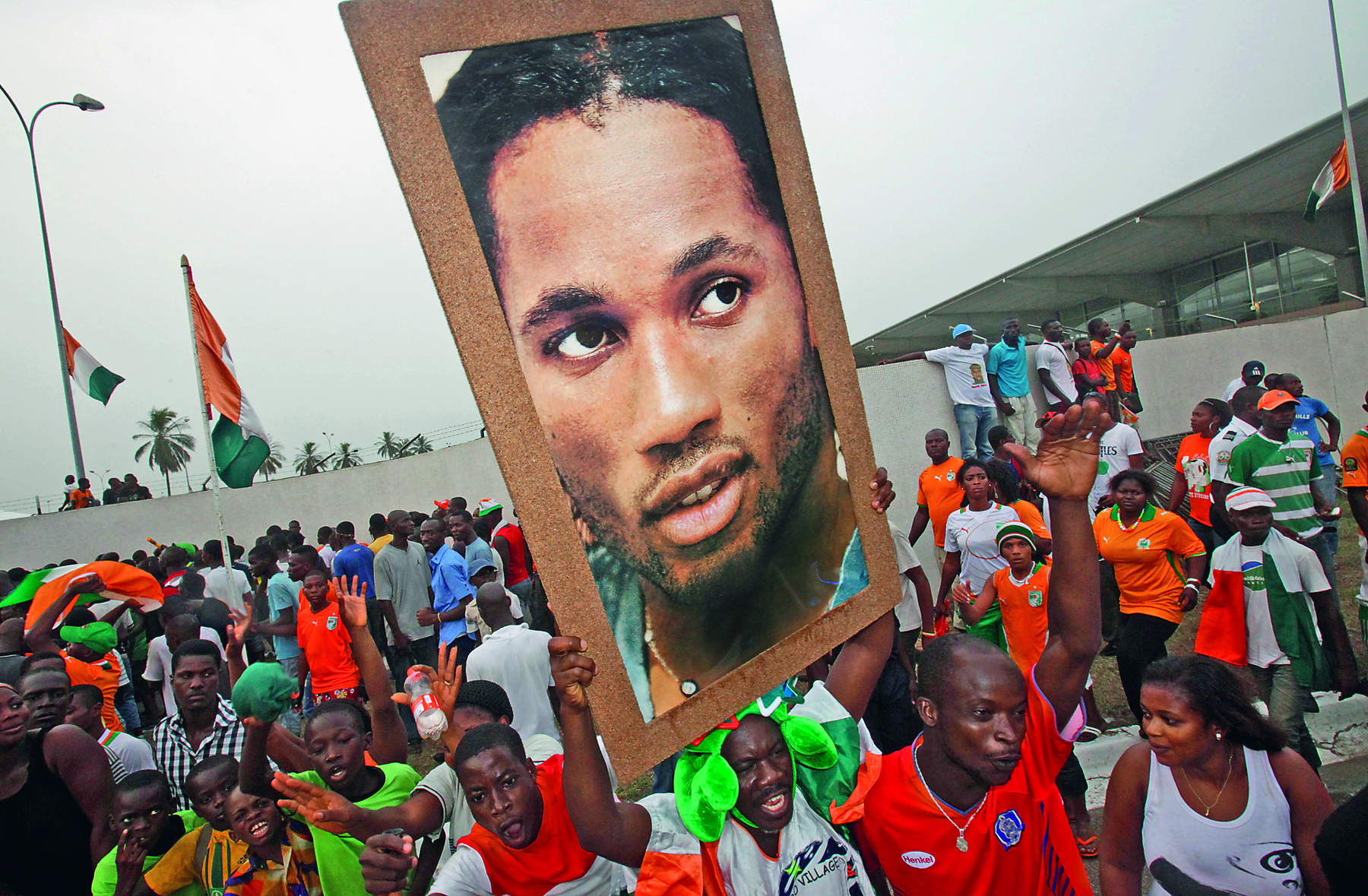 Fotografia. Pessoas negras aglomeradas na rua. Uma delas carrega um grande cartaz com a foto de Drogba, rapaz negro, de sobrancelhas curtas e lábios grossos.