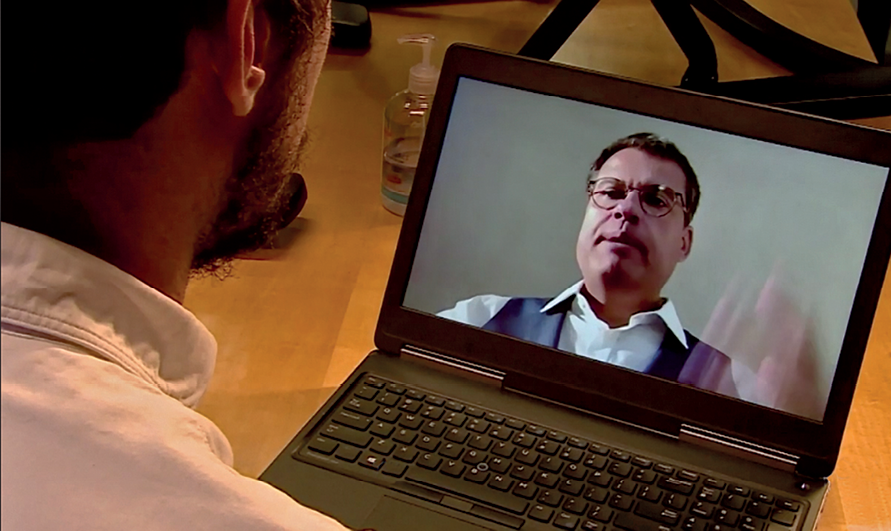 Fotografia. Rapaz em videochamada. À frente dele, um notebook aberto. Na tela, a imagem de Ricardo Falcão, homem de cabelo penteado de lado, rosto largo e óculos.