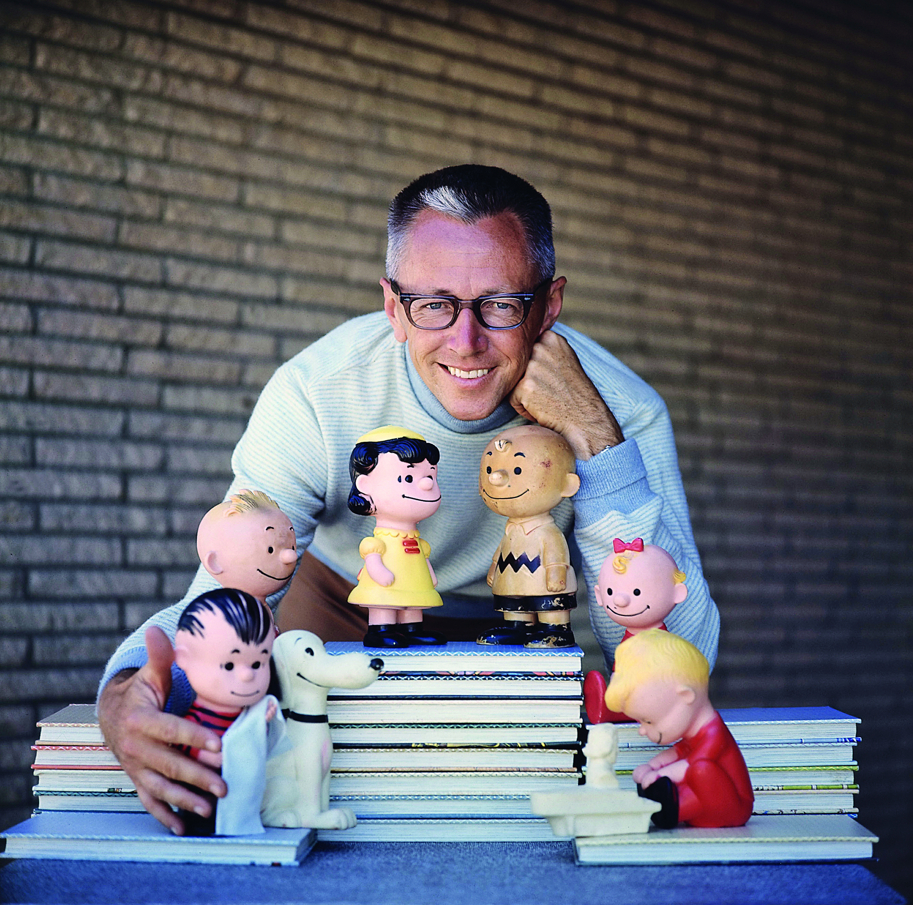 Fotografia. Charles M. Schulz. Homem de aproximadamente 50 anos, cabelo curto e grisalho. Usa óculos e armação escura. Está com o corpo inclinado para frente, com os braços ao redor de bonecos do desenho Charlie Brown.