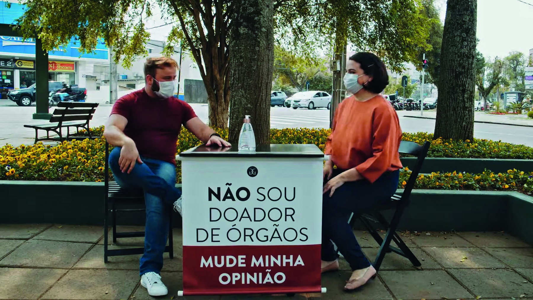 Fotografia. Um homem e uma mulher conversando em uma área aberta e arborizada. João é um homem branco de cabelo curto e pintado de loiro. Ele tem o corpo grande. Usa uma camiseta justa ao corpo. Estão sentados em cadeiras. Entre eles uma mesa quadrada. Na frente dela um banner pendurado que diz: NÃO SOU DOADOR DE ÓRGÃOS. MUDE MINHA OPINIÃO.