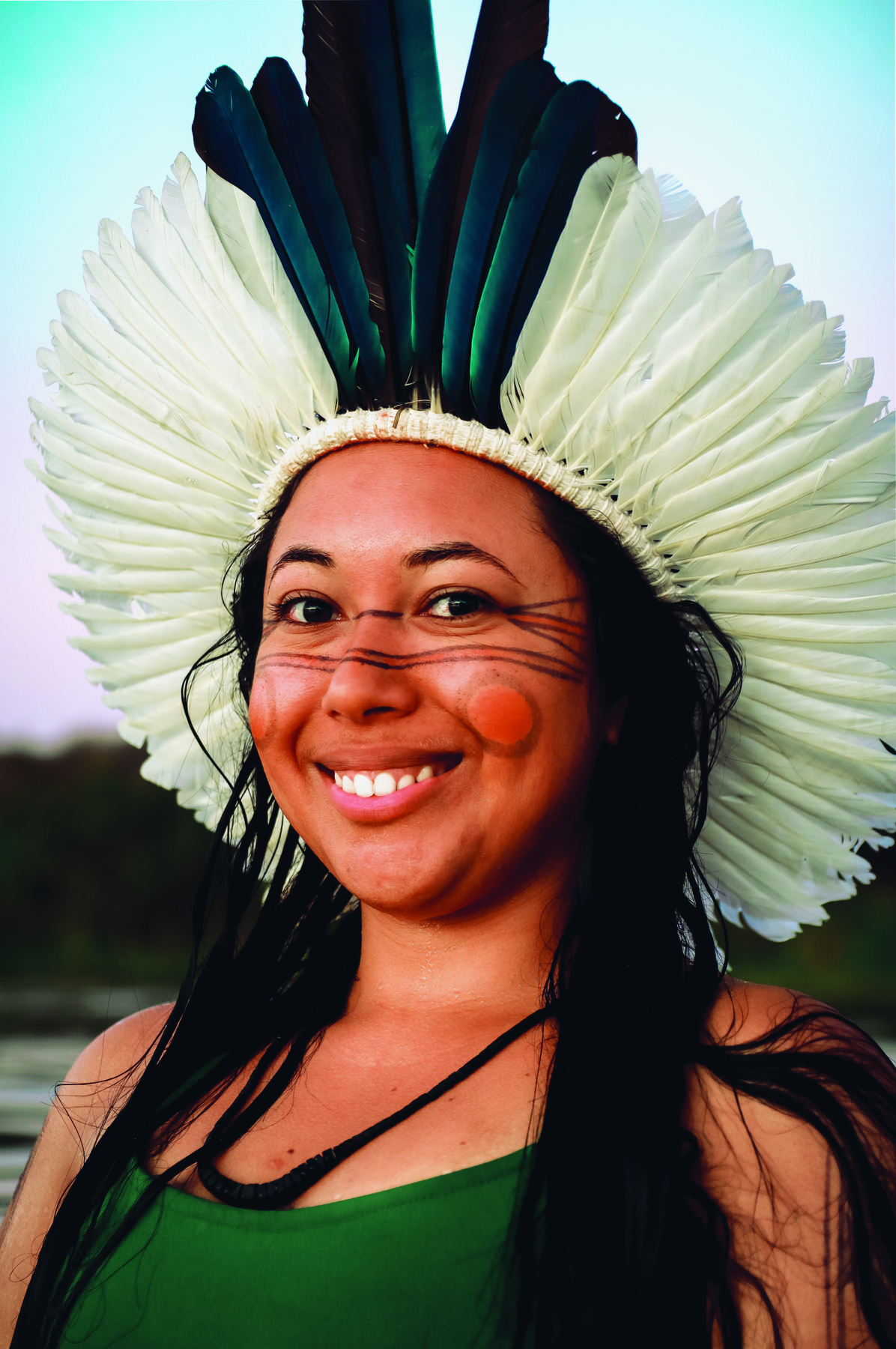 Fotografia. Hamangaí Pataxó, moça indígena, de pele morena, cabelo longo e escuro. Usa um cocar de penas brancas com algumas penas azuis no centro.