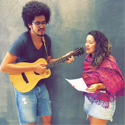 Fotografia. Jafté Salles, rapaz negro de cabelo Black Power. Usa óculos. Está em pé tocando um violão. Ao lado dele Larissa Muniz, moça de cabelo longo e encaracolado com as pontas douradas. Ela é mais baixa que ele. Usa um lenço rosa ao redor da parte superior do corpo. Ela tem uma folha nas mãos.