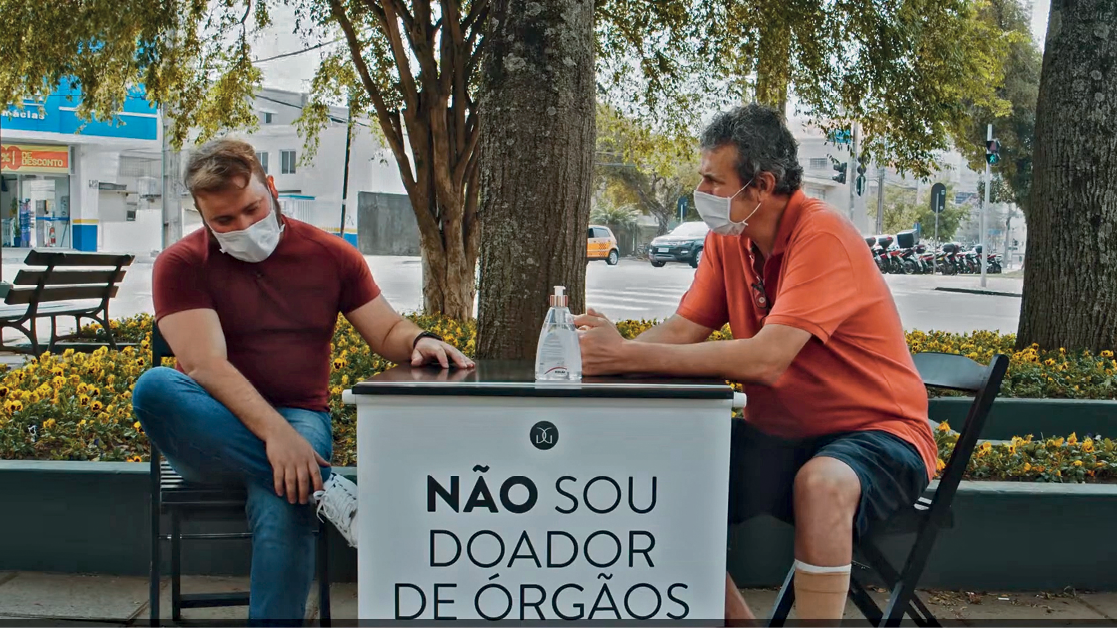 Fotografia. Conversa entre dois homens, João e Moacir. João sentado à esquerda e Moacir, um homem de cabelo grisalho e curto, usando máscara de proteção facial e camiseta laranja, está sentado à direita.