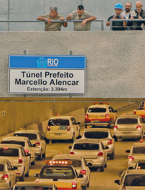 Fotografia. Homens em cima de um viaduto. Estão olhando para baixo, na direção de carros entrando no túnel. Acima da entrada para o túnel, vê-se uma placa azul e branca. A placa exibe o logotipo da Prefeitura do Rio de Janeiro, e o texto: Túnel Prefeito Marcello Alencar. Extensão: 3.394 metros. A palavra "extensão" está grafada com cê cedilha.