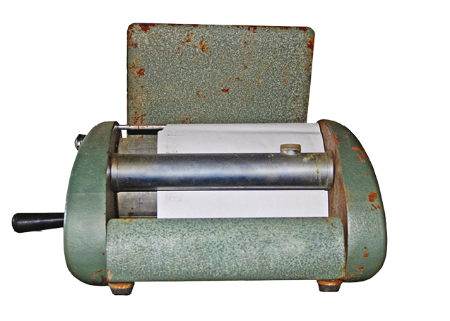 Fotografia. Mimeógrafo, aparelho de ferro largo com um cilindro no meio. A folha está presa em um cilindro encaixado em uma base com manivela. A folha copiada está na bandeja à frente do cilindro.