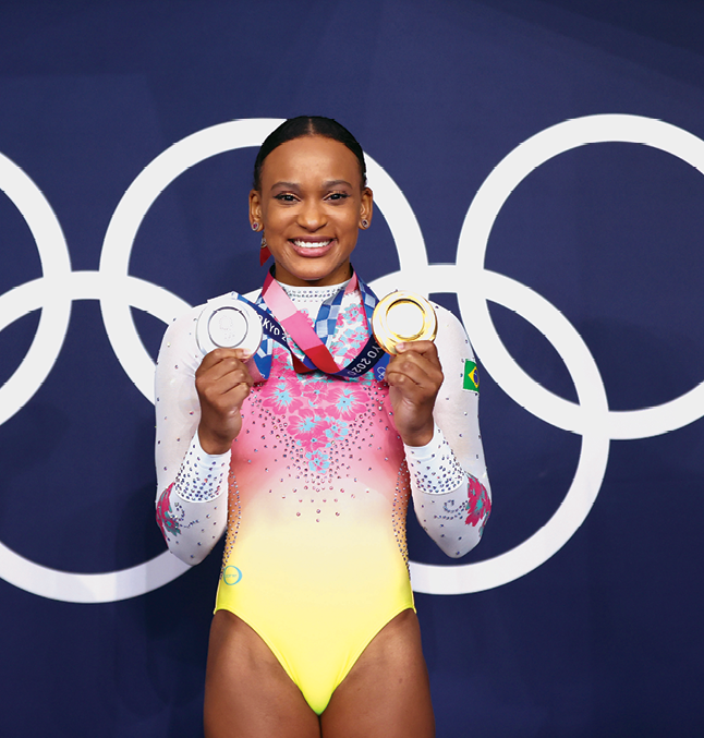 Fotografia. Rebeca Andrade, ginasta negra. Tem o cabelo penteado para trás, usa collant e exibe suas medalhas.