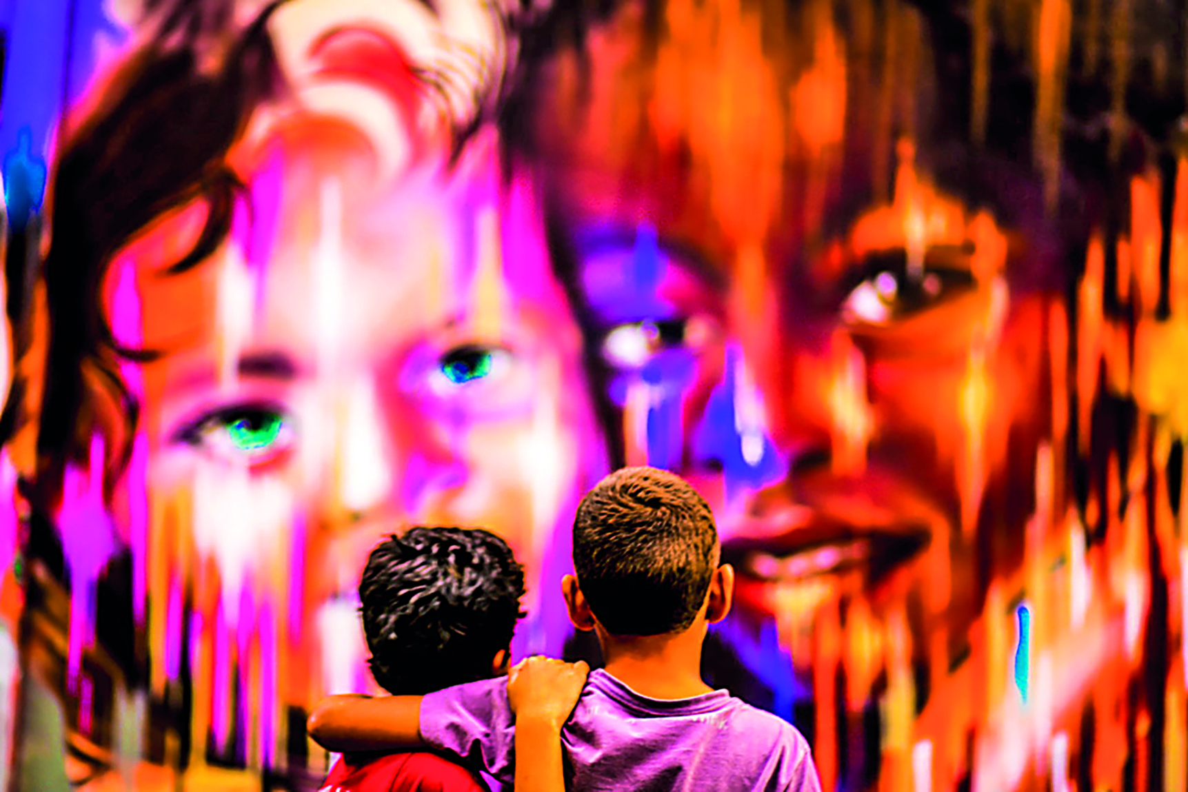 Fotografia. Dois meninos de costas. O da esquerda usa camiseta vermelha e está com a mão direita sobre o ombro do menino da direita de camiseta lilás e que está com o braço esquerda sobre os ombos do outro. Ao fundo, um muro com grafite colorido de dois rostos de meninos com partes escorrendo de tinta. À esquerda, rosto de menino de cabelos escuros e, à direita, menino de cabelos escuros.