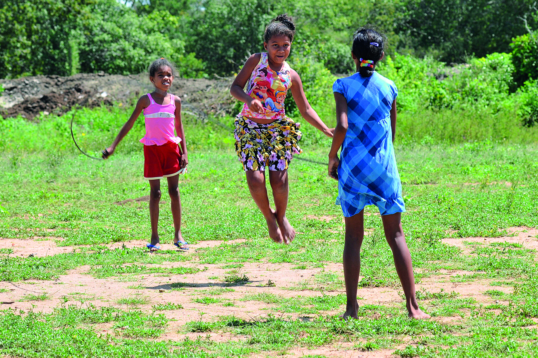 Fotografia. Em um local aberto, com grama e solo arenoso com três meninas brincando de pular corda. À frente, uma menina vista de costas, em pé, com vestido de mangas curtas em azul segurando a corda na mão direita. Ao centro, uma menina pulando a corda de blusa regata e saia, estampadas, com o corpo no ar e descalça. Ao fundo, menina de regata rosa e bermuda em vermelha, segurando a outra ponta da corda. Mais ao fundo, vegetação em verde.