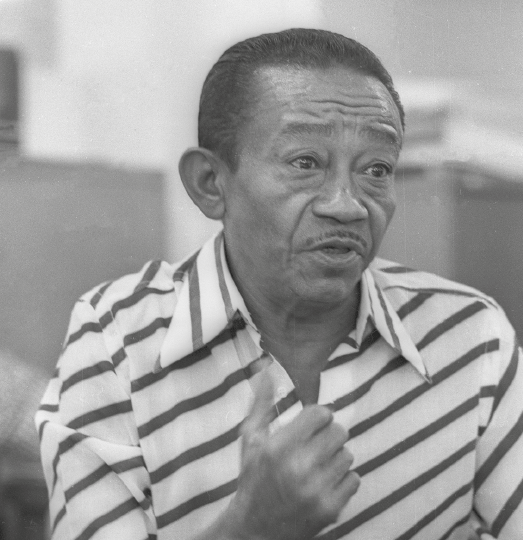 Fotografia em preto e branco. Jackson do Pandeiro, visto do busto para cima, negro, cabelos escuros, com camisa com listras.  Ele está com o cenho franzido e a boca aberta falando.