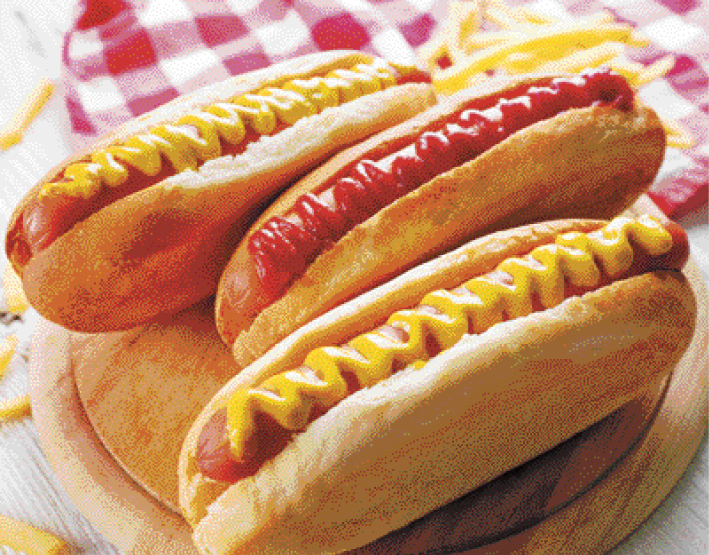 Fotografia. Vista de cima para baixo, sobre uma tábua de madeira redonda, três cachorros-quentes: pão comprido cortado no comprimento com uma salsicha no meio, dois com mostarda e um com ketchup por cima. Ao fundo, vista parcial de tecido em xadrez em branco e vermelho.