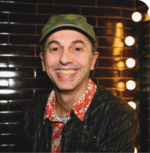 Fotografia. Músico Zeca Baleiro, homem visto do busto para cima, olhando para frente sorrindo. Ele tem cabelos escuros para trás e usa boné verde sobre a cabeça, nariz fino e blusa de gola em vermelho com estampas em bege e jaqueta preta por cima. Ele olha para frente sorrindo. Ao fundo, local com luzes verticais à direita.