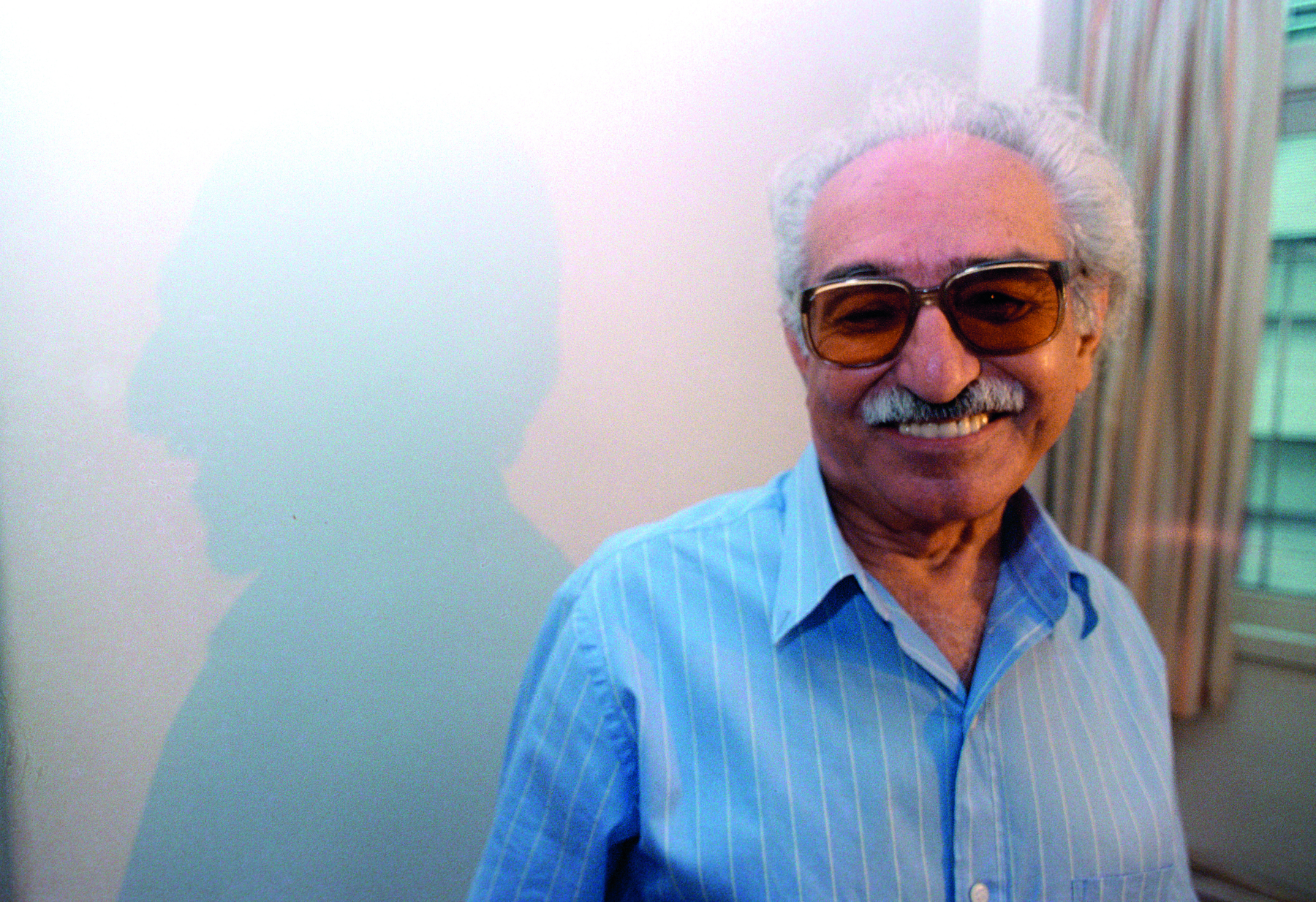 Fotografia. Poeta Manoel de Barros, homem visto dos ombros para cima,  cabelos e bigode grisalhos, com par de óculos de sol em marrom. Ele olha para frente sorrindo, usando camisa de gola em azul-claro. Ao fundo, local com parede branca e partes em marrom na vertical.