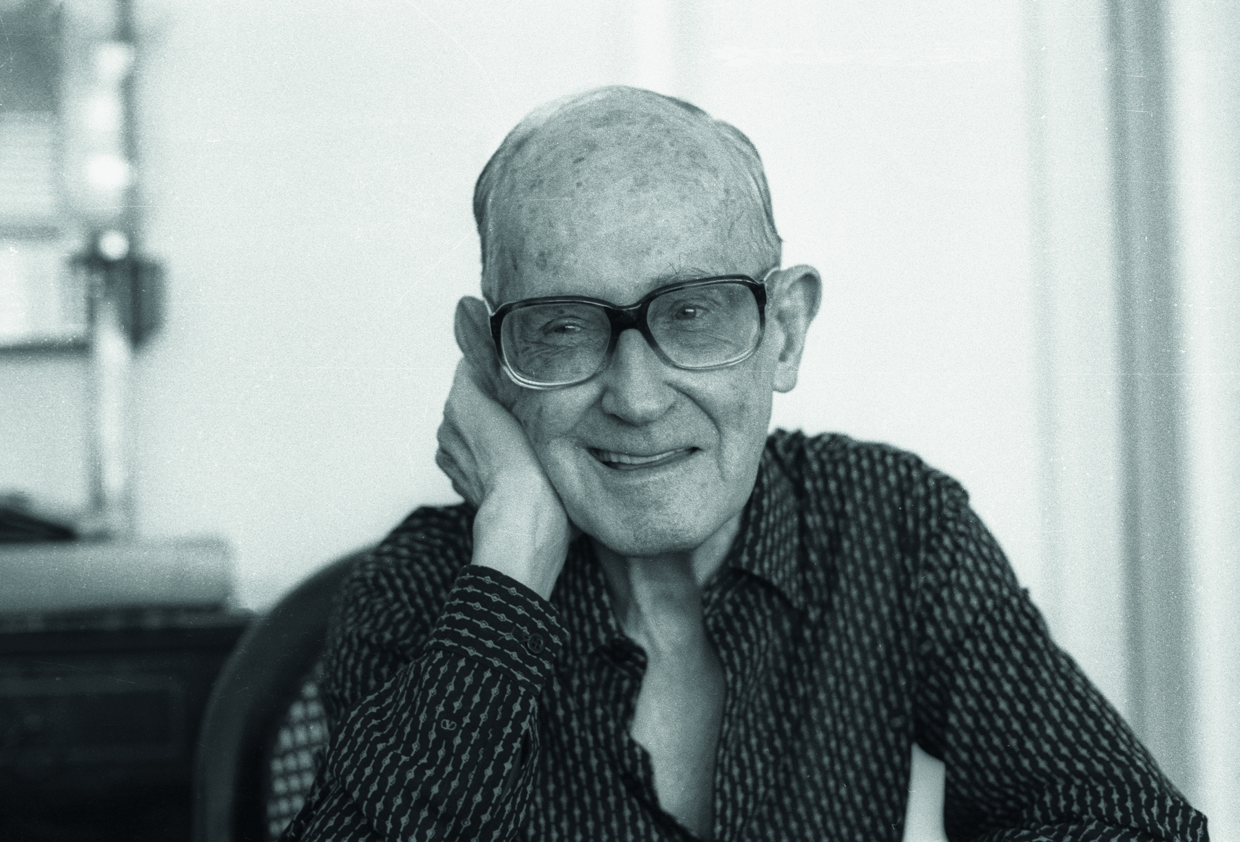 Fotografia em preto e branco. Imagem do poeta Carlos Drummond de Andrade. Homem senhor visto do busto para cima, branco, careca, com par de óculos de grau de formato quadrado, com camisa de mangas compridas escuro com pontos claros e braço direito apoiando a cabeça. Ele olha para frente sorrindo.