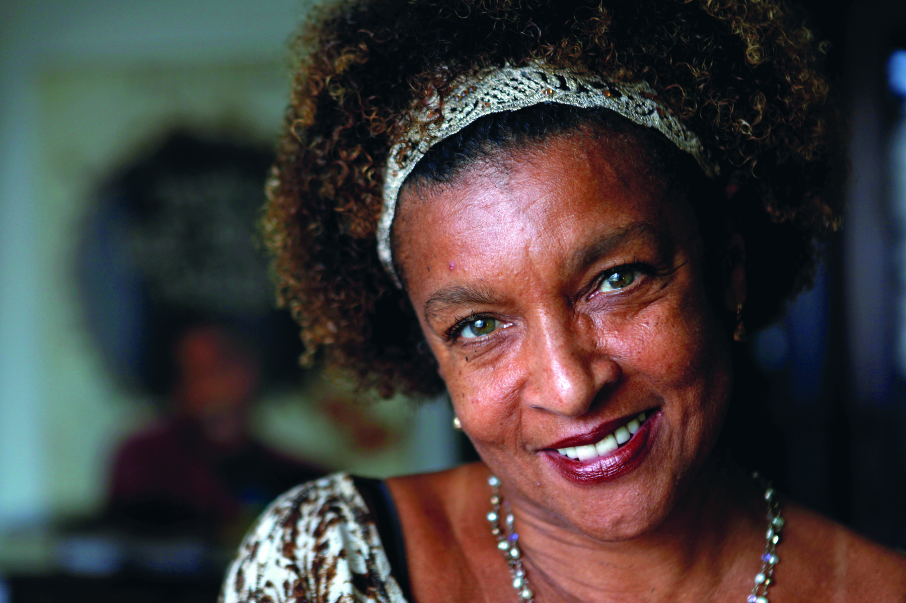 Fotografia. Artista Elisa Lucinda, mulher negra, vista do busto para cima, cabelos encaracolados  marrom, com uma faixa sobre os cabelos em cinza. Ela usa batom vermelho-escuro, olha para frente sorrindo com a cabeça levemente inclinada para direita, colar no pescoço e roupa em branco e marrom.