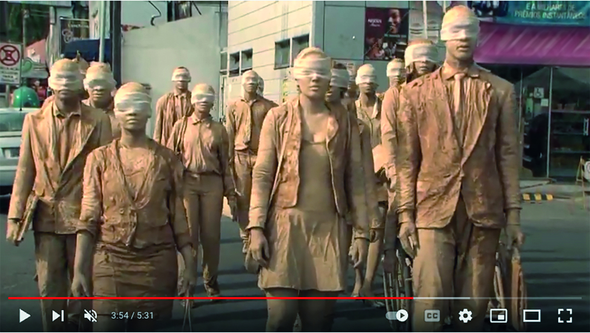 Captura de vídeo. Grupo doze pessoas visíveis na imagem, homens e mulheres caminhando para frente, levando algo na mão. Todos estão com um tecido branco tampando os olhos, homens vestidos de terno em marrom-claro e mulheres com a mesma cor de roupa, algumas com saia. Em segundo plano, vista parcial de prédios de cidade. Na parte inferior, fio vermelho e branco e ícones de canal de vídeos.