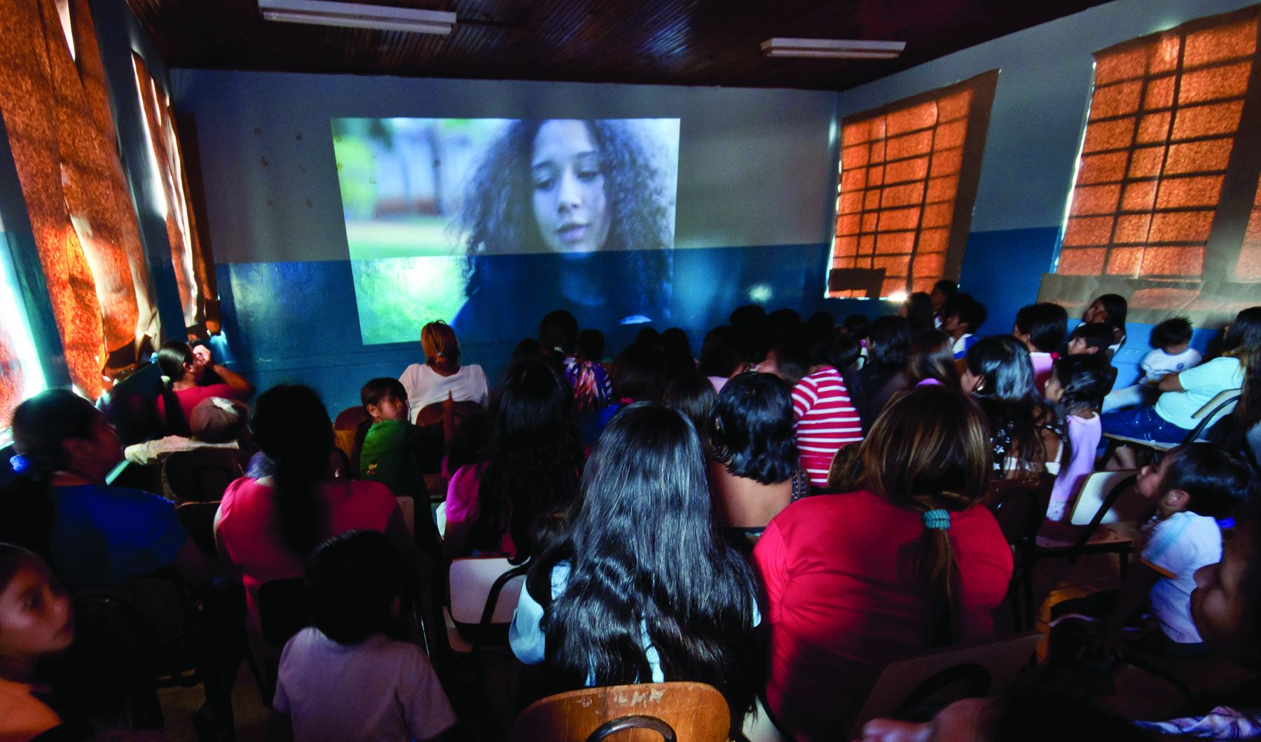 Fotografia. Vista geral de uma sala de aula com paredes brancas da metade para cima e azul da metade para baixo, nela há carteiras escolares com pessoas de várias idades e características físicas sentadas de costa. Ao fundo, na parede, projeção de vídeo em que aparece mulher de cabelos compridos. Nas laterais janelas fechadas com cortinas para deixar o ambiente mais escuro.