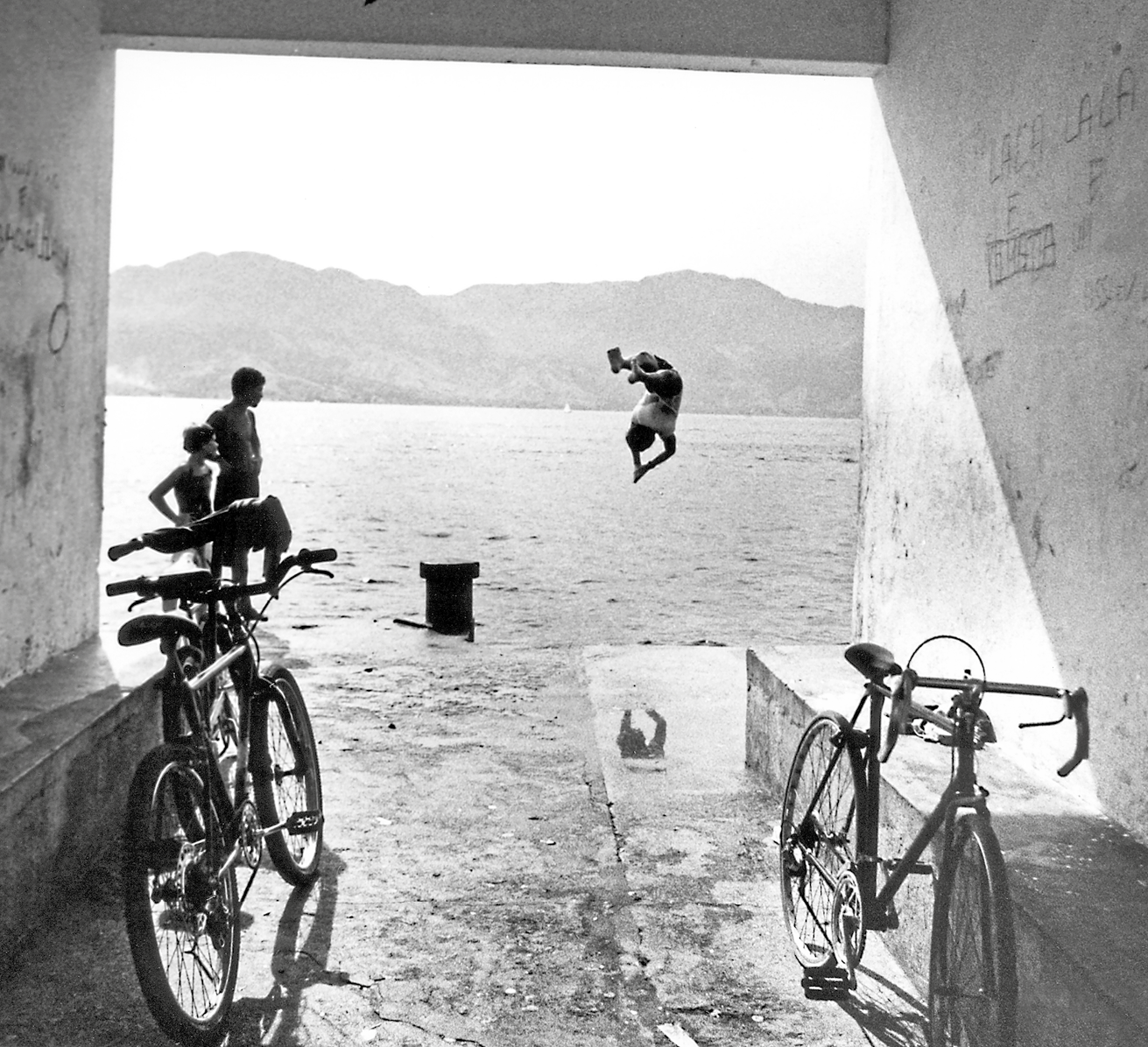 Fotografia em preto e branco. Local com paredes à esquerda e à direita. E muro baixo em ambos os lados com bicicletas encostadas na esquerda e na direita. Mais ao fundo, à esquerda, duas crianças, vistas de costas, perto uma da outra. À frente, uma criança com o corpo no ar, com os braços apontados para baixo para em direção a local com o mar. Em segundo plano, morros escuros e céu claro.