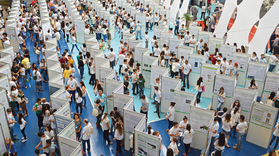 Fotografia. Vista do alto de salão com chão azul e com painéis brancos, em zigue-zague dividindo o espaço em vários corredores e várias pessoas em pé pelo ambiente.