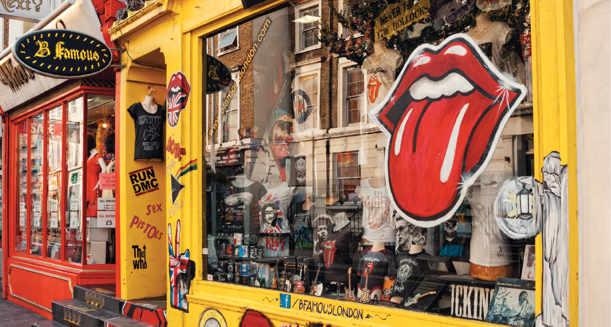 Fotografia. Fachada de lojas em Londres. À esquerda uma loja com detalhes em vermelho e vitrines; à direita uma loja amarela, com vitrine com artigos relacionados à música, como miniaturas e camisetas, na vitrine um adesivo à direita de uma ilustração de uma boca com a língua para fora, símbolo da banda inglesa, Rolling Stones.