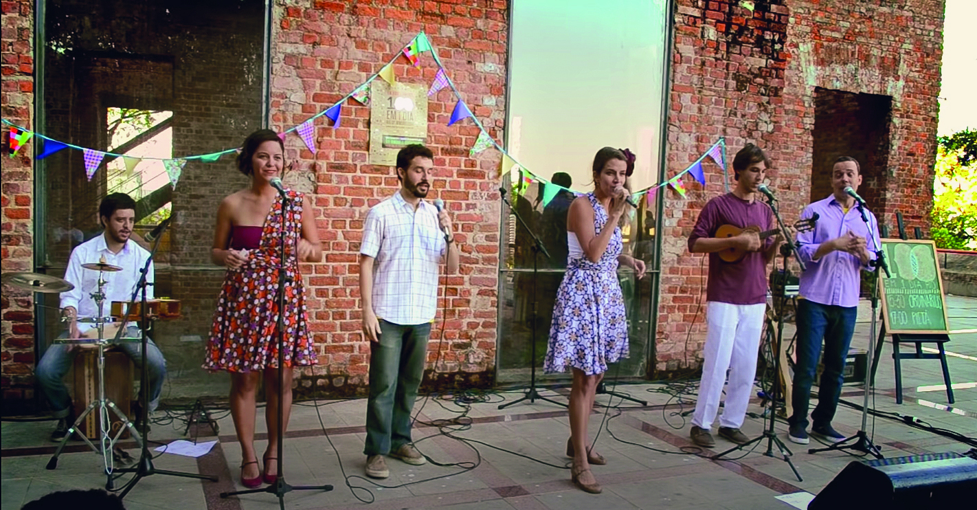 Fotografia. Local aberto, com piso cinza e parede de tijolos marrom ao fundo, seis pessoas cantando e tocando instrumentos. Da esquerda para direita: baterista de cabelos escuros, camisa branca, calça cinza e par de sapatos pretos. À direita, duas mulheres de vestidos estampados, cantando de frente para microfones. Entre elas, um homem cantando, ele tem  cabelos e barba escuro, camisa branca, calça cinza e sapatos marrons. Ele segura na mão direita, um microfone cinza. Na ponta da direita, outros dois homens cantando de frente para microfones. Um deles tem cabelos castanhos e usa blusa vinho, calça branca e sapatos marrons e tocando um cavaquinho. Ao fundo, sobre as paredes, uma fita na horizontal com muitas bandeirinhas coloridas.