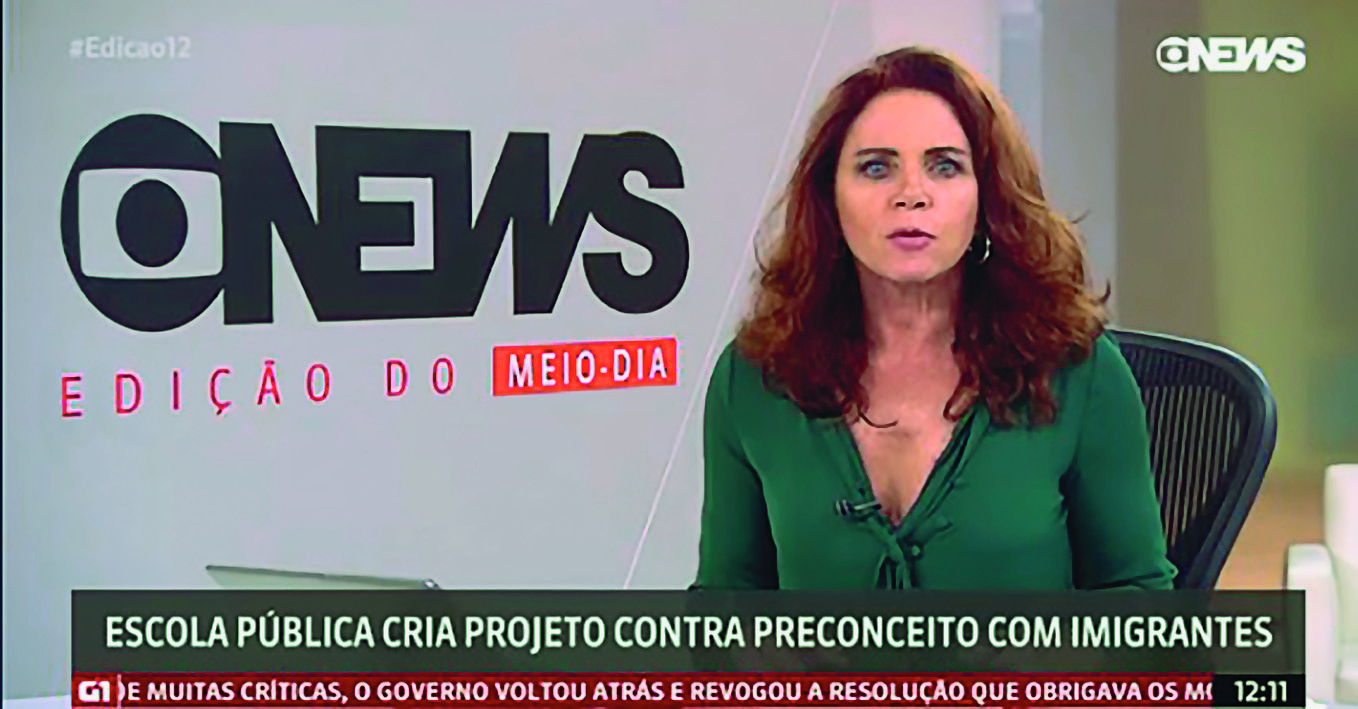 Captura de tela de TV. Mulher apresentando um telejornal. À direita, mulher sentada sobre cadeira preta. Ela tem  cabelos ruivos penteados para o lado, veste blusa verde de mangas compridas. À esquerda, parede branca com texto: Globonews EDIÇÃO DO MEIO-DIA. Abaixo, em fundo cinza, legenda: ESCOLA PÚBLICA CRIA PROJETO CONTRA PRECONCEITO COM IMIGRANTES. Abaixo em fundo vermelho, manchetes: G1. E MUITAS CRÍTICAS, O GOVERNO VOLTOU ATRÁS E REVOGOU A RESOLUÇÃO QUE OBRIGAVA OS... Na ponta superior, à direita, logotipo: Globonews.