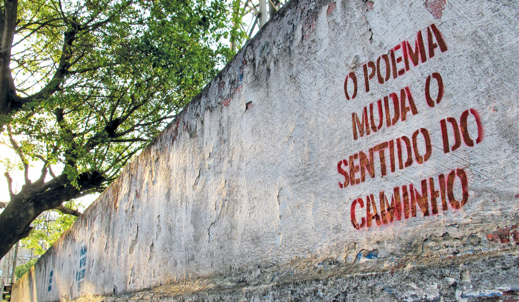 Fotografia. Um muro branco cruzando a foto diagonalmente da parte inferior esquerda para a parte superior direita. À direita dele, texto em vermelho pintado na parede: O POEMA MUDA O SENTIDO DO CAMINHO. Na parte superior esquerda, troncos e galhos marrons e folhas verdes. Por trás das árvores,, céu em azul-claro com luz solar.