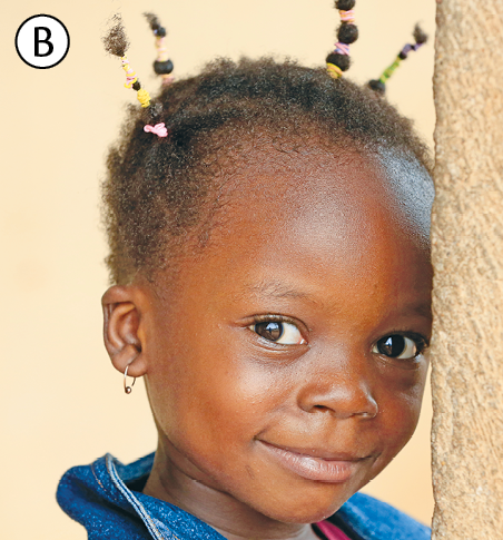 Fotografia B. Busto de uma menina de cabelos crespos e ralos, com algumas tranças finas. Ela usa brincos pequenos de argola e veste blusa com gola azul. A menina olha para frente, sorrindo.