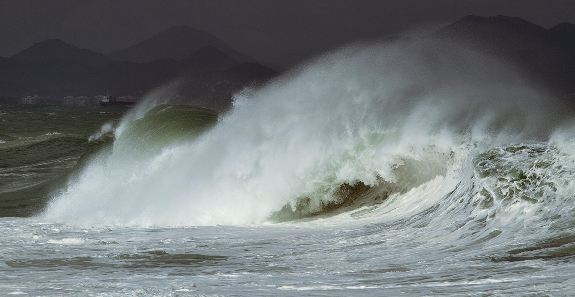 Fotografia. Destaque para as ondas do mar se quebrando. O mar está com tons bem escuros de verde e branco na parte das ondas. Ao fundo, céu escuro e morros.