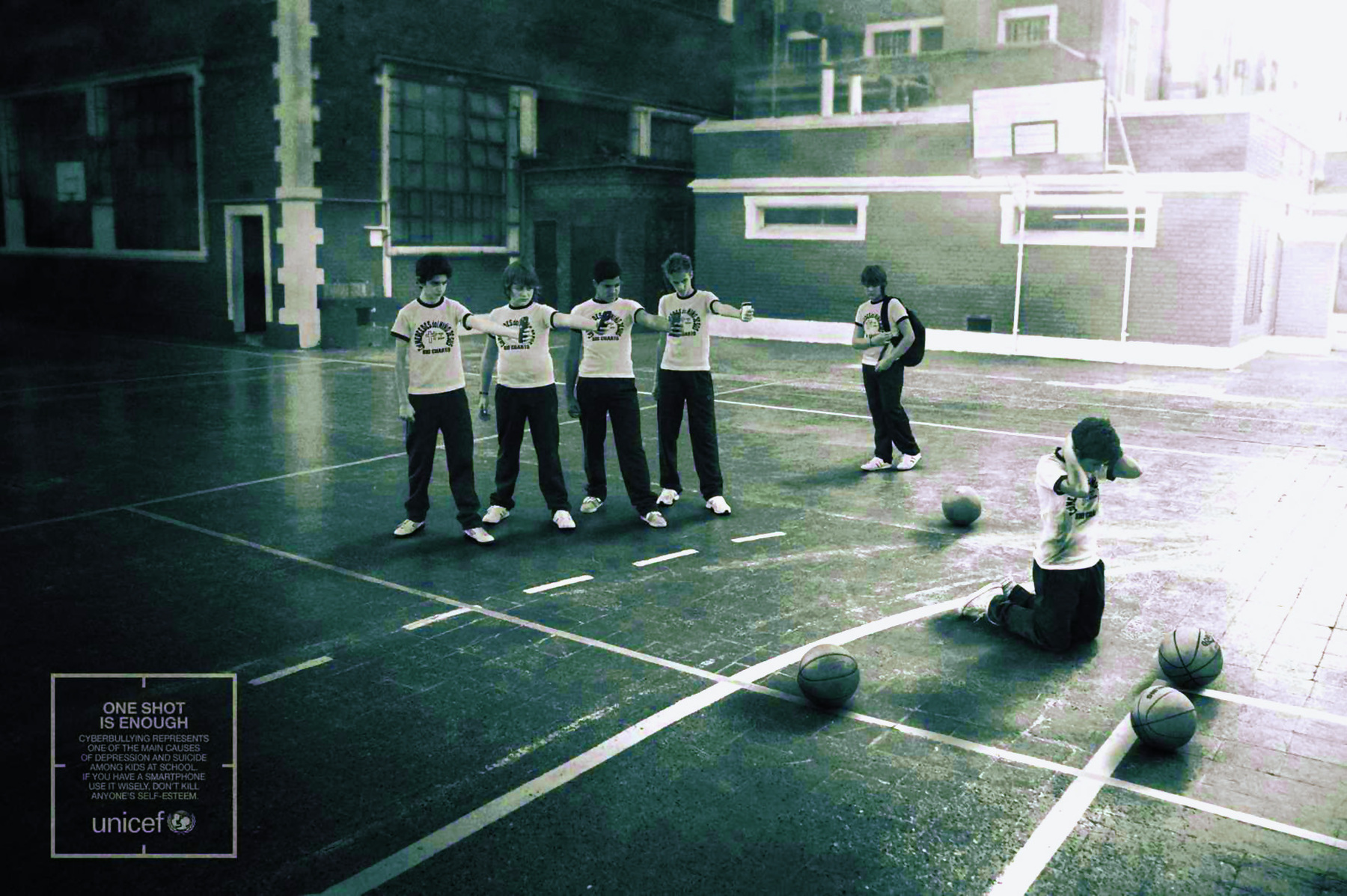 Fotografia em preto e branco. A fotografia mostra um grupo de crianças em uma quadra esportiva escolar. As crianças estão uniformizadas com camisetas brancas com estampa ao centro do peito, calças pretas e tênis claro. No centro da imagem, quatro crianças estão em pé com os braços esquerdos estendidos para frente e os punhos cerrados. Elas olham para uma criança ajoelhada mais à frente, a qual está com as mãos para trás da nuca e com o pescoço levemente inclinado para frente. Ao redor dessa criança, há quatro bolas de basquete espalhadas. Ao lado do grupo de quatro crianças em pé, há uma que se aproxima do grupo, caminhando. Ao fundo, há um cesto de basquete e paredes com janelas de diversos tamanho. Na parte inferior esquerda, em tamanho pequeno, há um texto em inglês e a logo da Unicef.